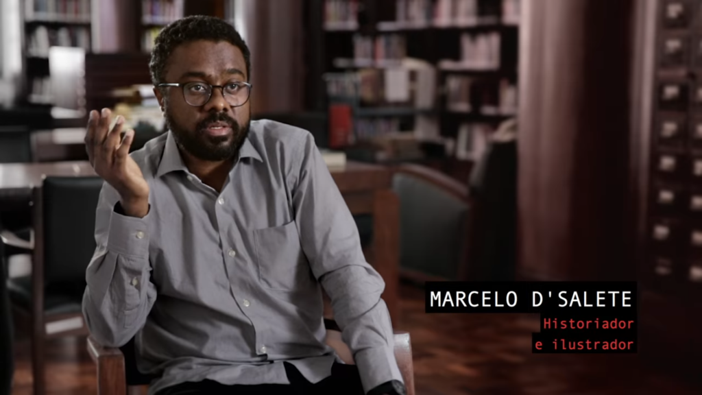 Marcelo D'Salete está numa biblioteca ou livraria. Usando camisa social de manga longa e cinza. Ele usa óculos também. Está falando algo. Está com a mão direita levantada na altura do rosto.