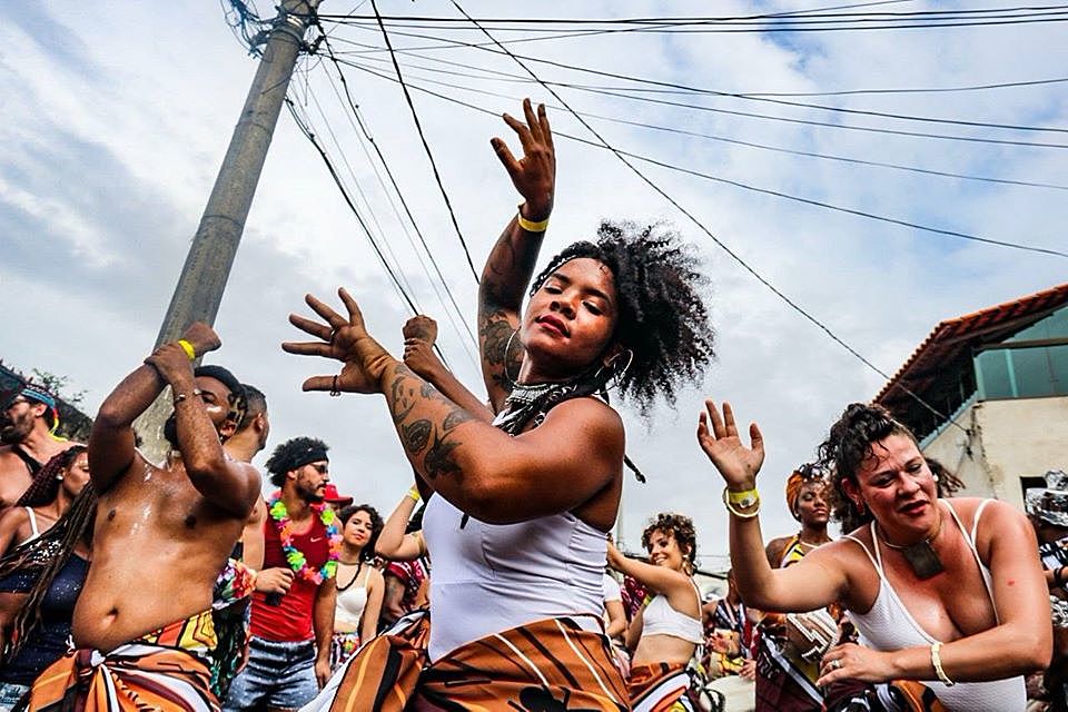 Foto de uma festa de rua. Pessoas (em sua maioria negras)  dançam alegremente. Usam roupas leves , ou não usam a arte de cima.
