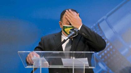 O General Pazuello está em pé atrás do púlpito, usando mascara (estampada com a bandeira do Brasil), e passando a mão na testa. 
