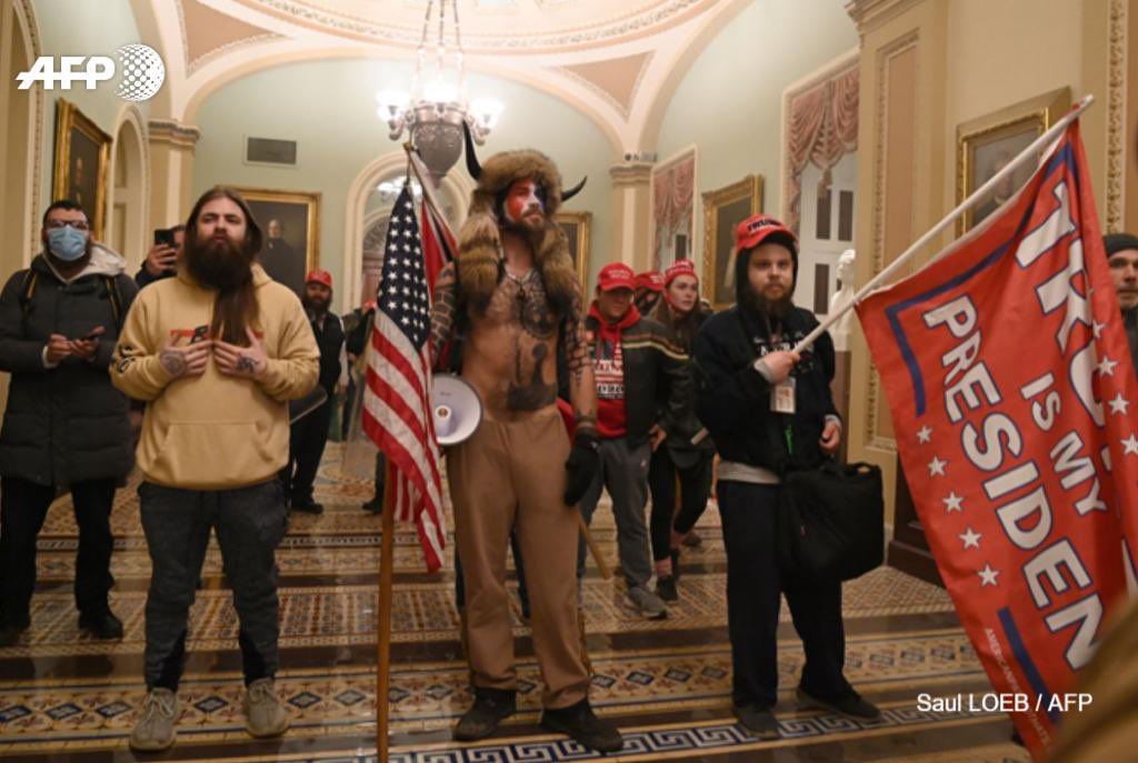 Terroristas supremacistas brancos, portando bandeiras escritas "Trump is my president". Eles usam bonés MAGA. O chamado "Shaman do QAnom" está no centro da foto.
