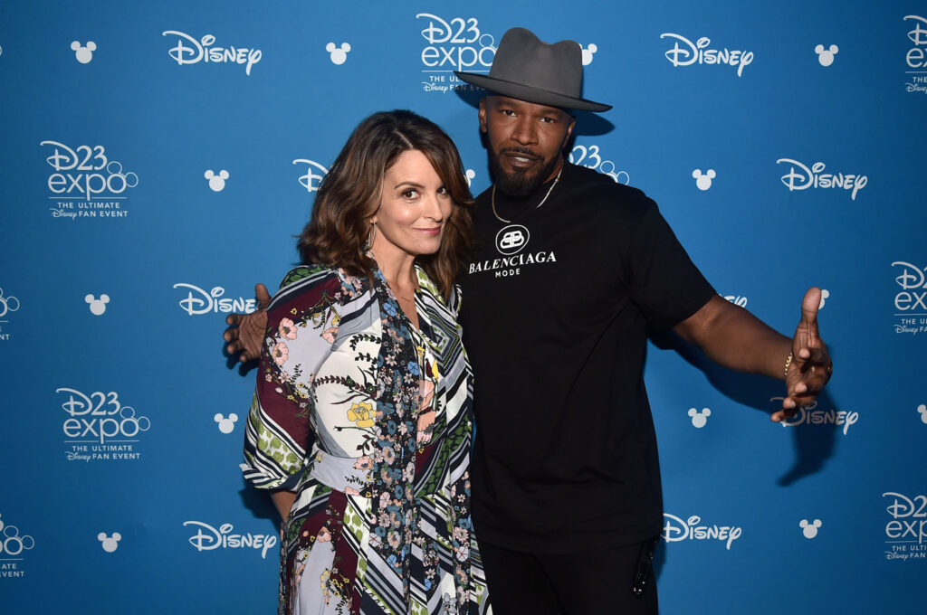 De frente à um banner com logos da Disney, o símbolo do Mickey, e a logo D23 Expo, estão Tina Fey e Jamie Foxx. Jamie está passando o braço por trás de Tina, mas não está abraçando ela. Está meio com os braços abertos e mãos espalmadas, como quem está declamando um rap.