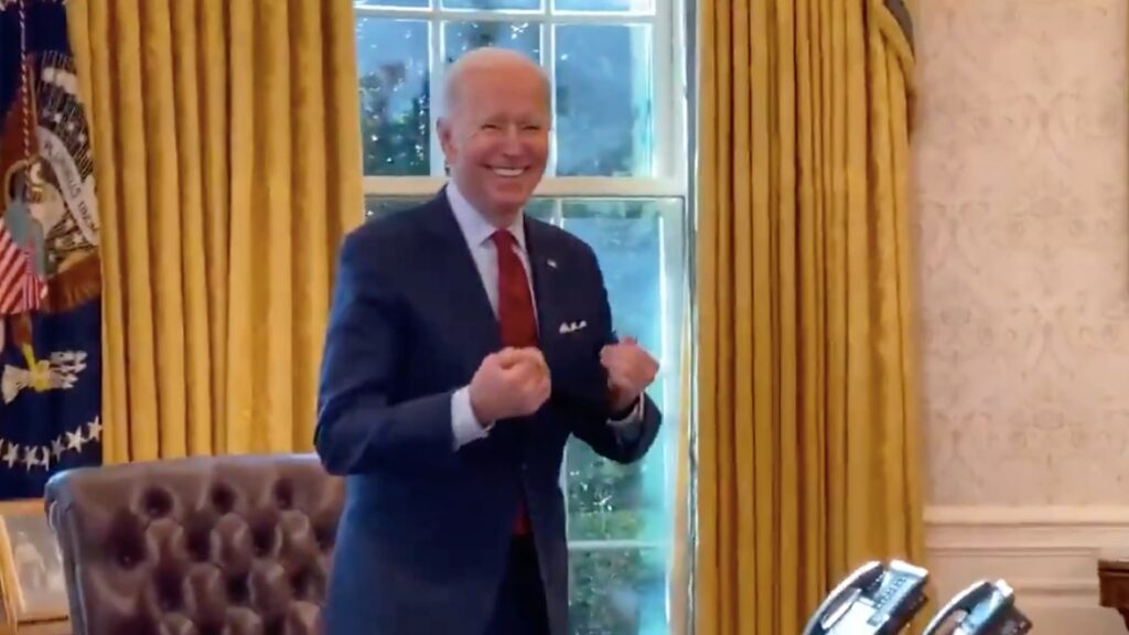 No salão oval, em pé atrás da mesa presidencial, Joe Biden está sorrindo, e com as mãos "em forma de coxinha" enquanto ri. 