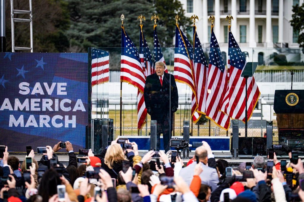 Num comício, o ex-presidente Donald Trump está falando com a platéia. No público, vemos diversos celulares filmando e tirando fotos. Muitas bandeiras dos EUA estão atrás de Trump, e ele está do lado de um painel onde se lê "Save America March".