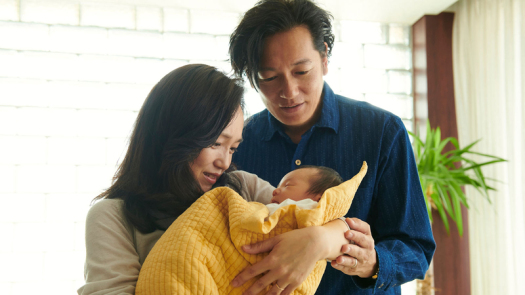 Imagem de Mães de Verdade. O casal Satoko e Kiyokazu está abraçado, observando um bebê de colo.