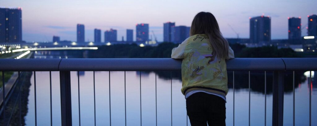 Imagem de Mães de Verdade.
Uma moça, de costas, olhando a cidade. Ela está sobre uma ponte, que está acima de um rio.