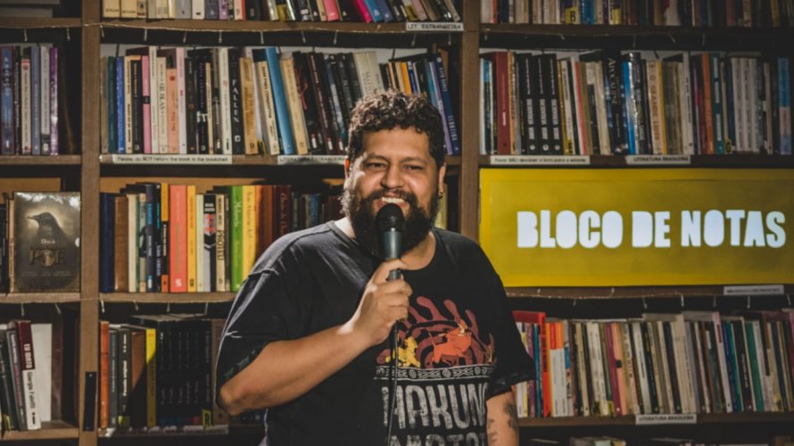 Foto de Matheus Buente em frente à uma estante de livros, em seu show de stand-up. Ao fundo, na direita, há um letreiro escrito "Bloco de Notas"