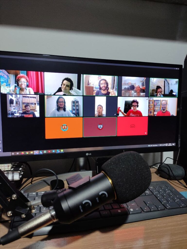 em primeiro plano, microfone usado para gravação de podcast posicionado em cima de um teclado. Ao fundo, o monitor de computador mostrando a tela do Discord com os participantes da gravação do último Maratona.