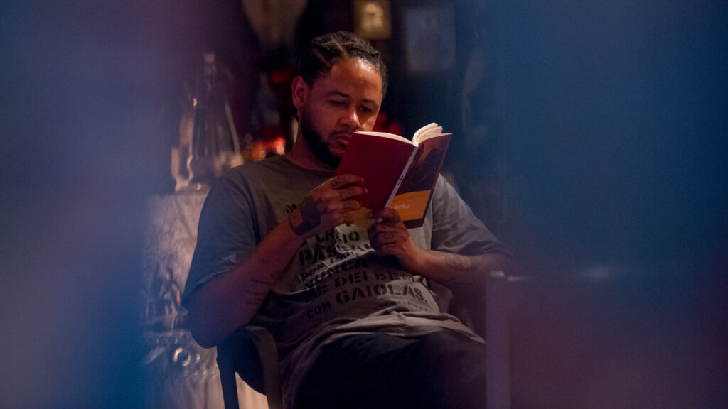 Cena de Medida Provisória. Berto (Emicida), dentro do Afro Bunker, lendo um livro (não dá pra identificar)