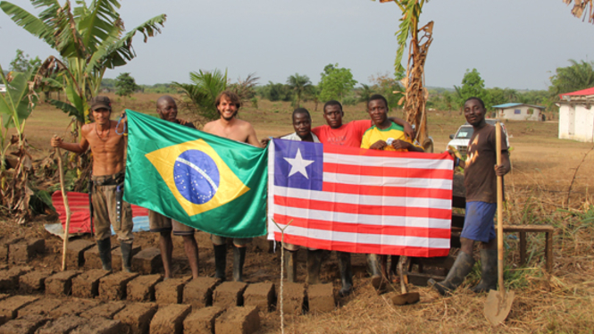 Homens brasileiros e liberianos em pé, atrás de duas bandeiras: na esquerda, a do Brasil, na direita a da Libéria. As pessoas estão numa área rural. Ao fundo pode-se ver uma planície, com vegetação ao fundo, e pequenas casas brancas ao fundo, na direita.