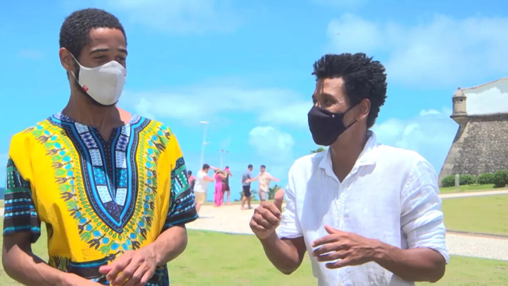 Alfred Enoch e Audri Anunciação, no Faro da Barra (Salvador/Ba). É um dia ensolarado, e existem pessoas ao fundo. Podemos ver o Farol ao fundo, na direita.