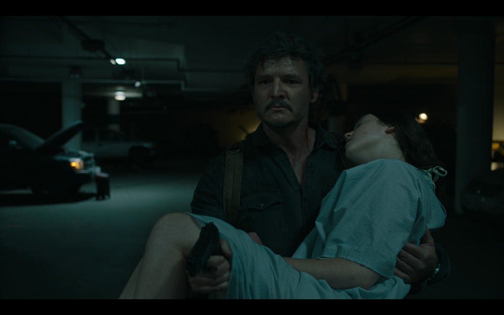 Cena de The Last of Us, onde Joel carrega Ellie nos braços, e Empunha uma arma com a mão direita. Ellie está com roupa de hospital. Eles estão dentro de um estacionamento.