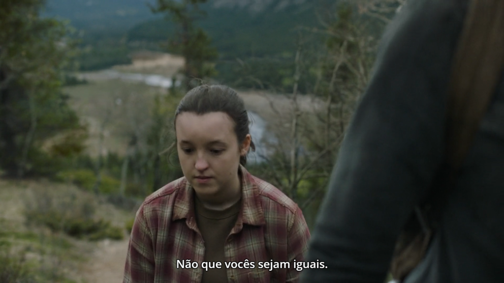 Cena de The Last of Us. A câmera mostra o braço de Joel em primeiro plano mas foa em Ellie, logo atrás. Ela está olhando para a trilha, mas sem muita animação. Atrás dela, uma paisagem natural, a planice abaixo da montanha que estão subindo. Na legenda, "Não que vocês sejam iguais."